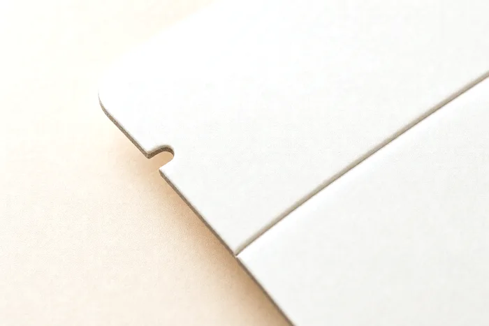 Close-up of a cut edge and scored crease on a blank carton corner, with a steel-rule cutting die board out of focus behind.
